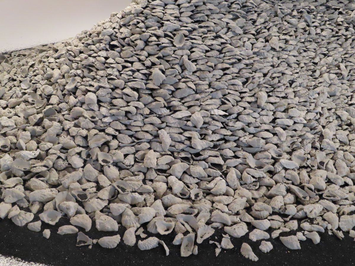 小山丘是用一片片灰白色、形如牡蠣殼的物件層層堆疊而成，代表原本存在這片土地上的貝塚，其間灑落的是帶有玻璃光澤的黑色銅渣